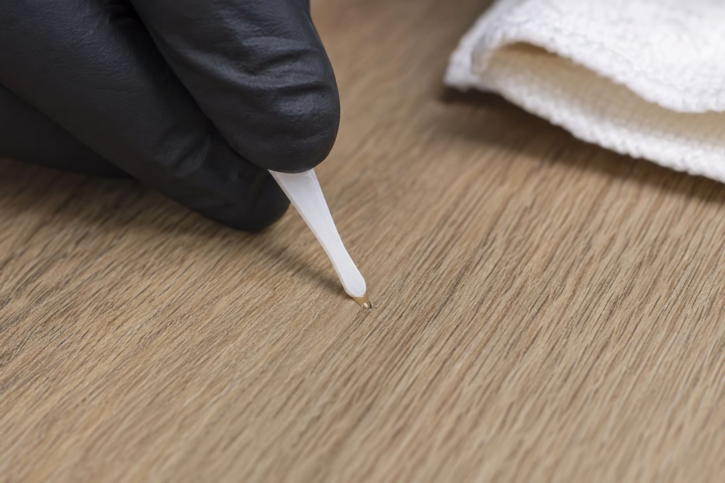 A gloved hand fills a small stable gouge in vinyl plank flooring with matching repair compound.