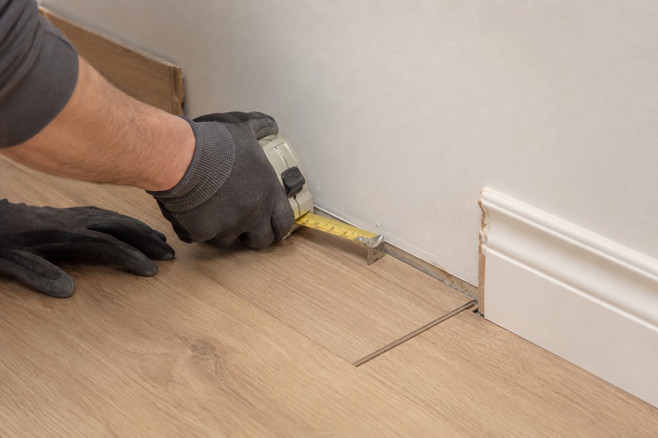 Flooring installer measures the perimeter clearance of click-lock vinyl planks beside a wall with a section of baseboard removed.